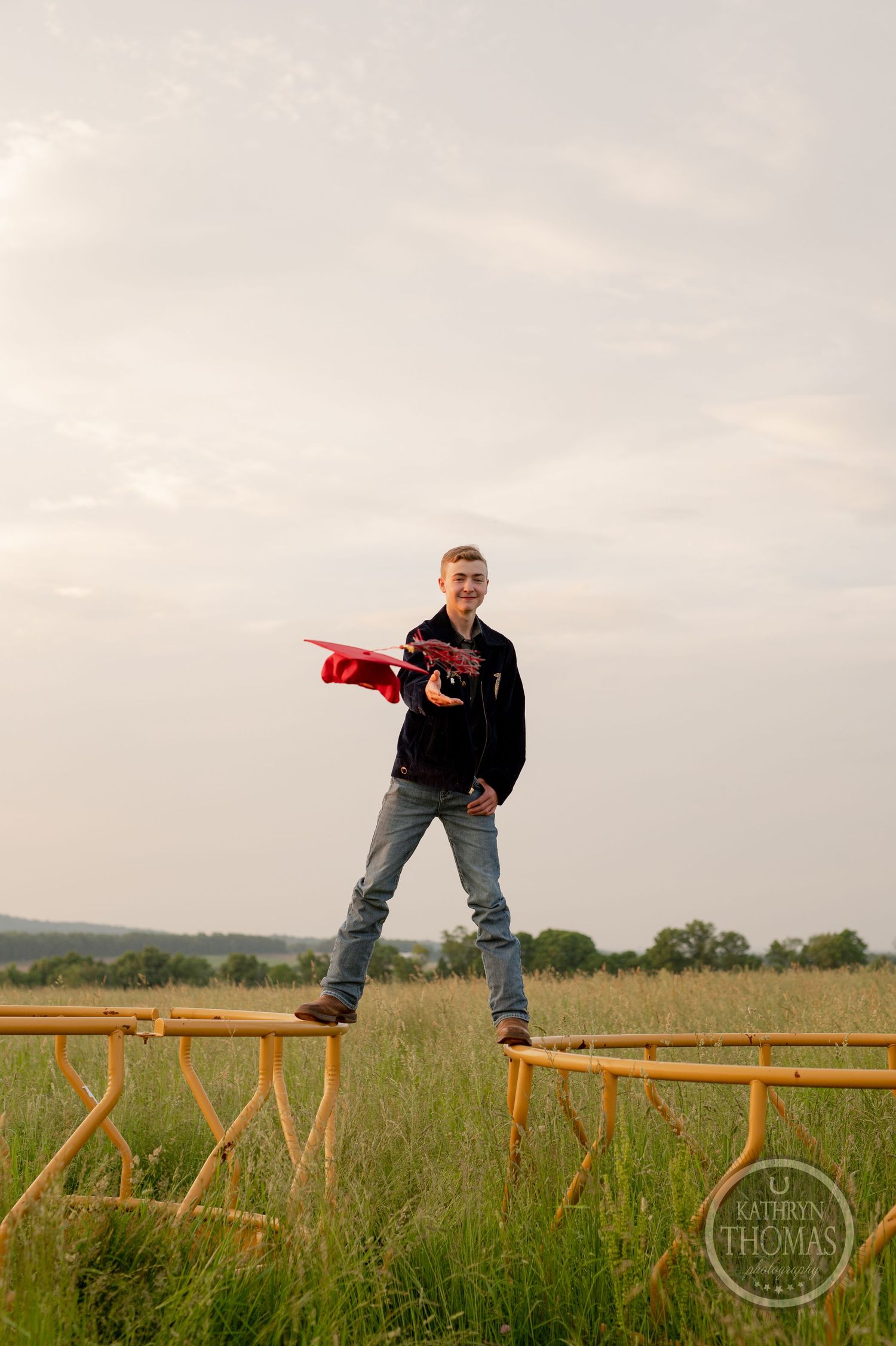 Student balancing on wooden fence holds red graduation cap against cloudy sky.