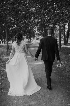 Newlyweds walking hand in hand down a tree-lined path in black and white.