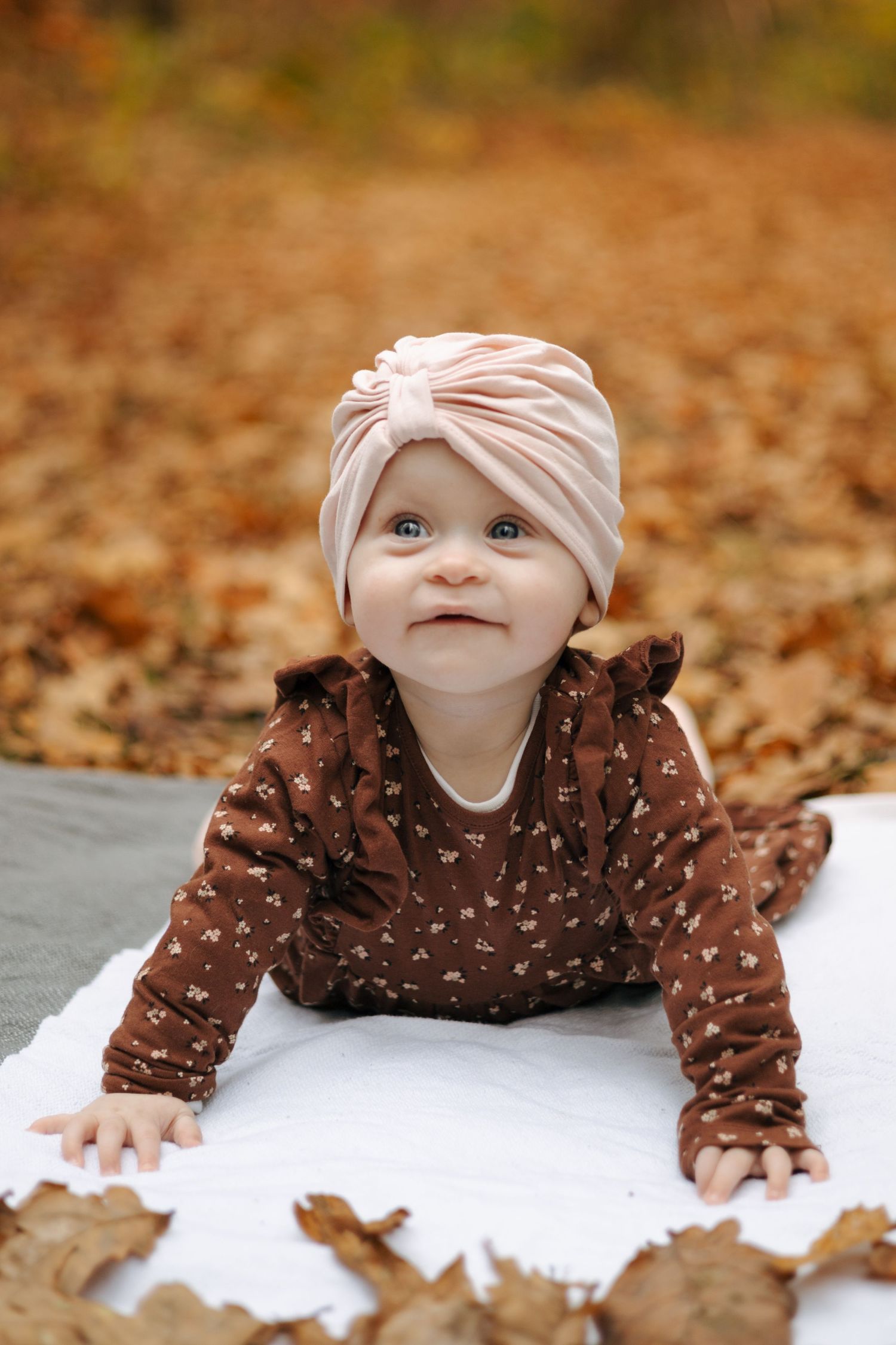 Baby wearing pink turban and brown polka dot outfit lying on blanket among fall leaves.