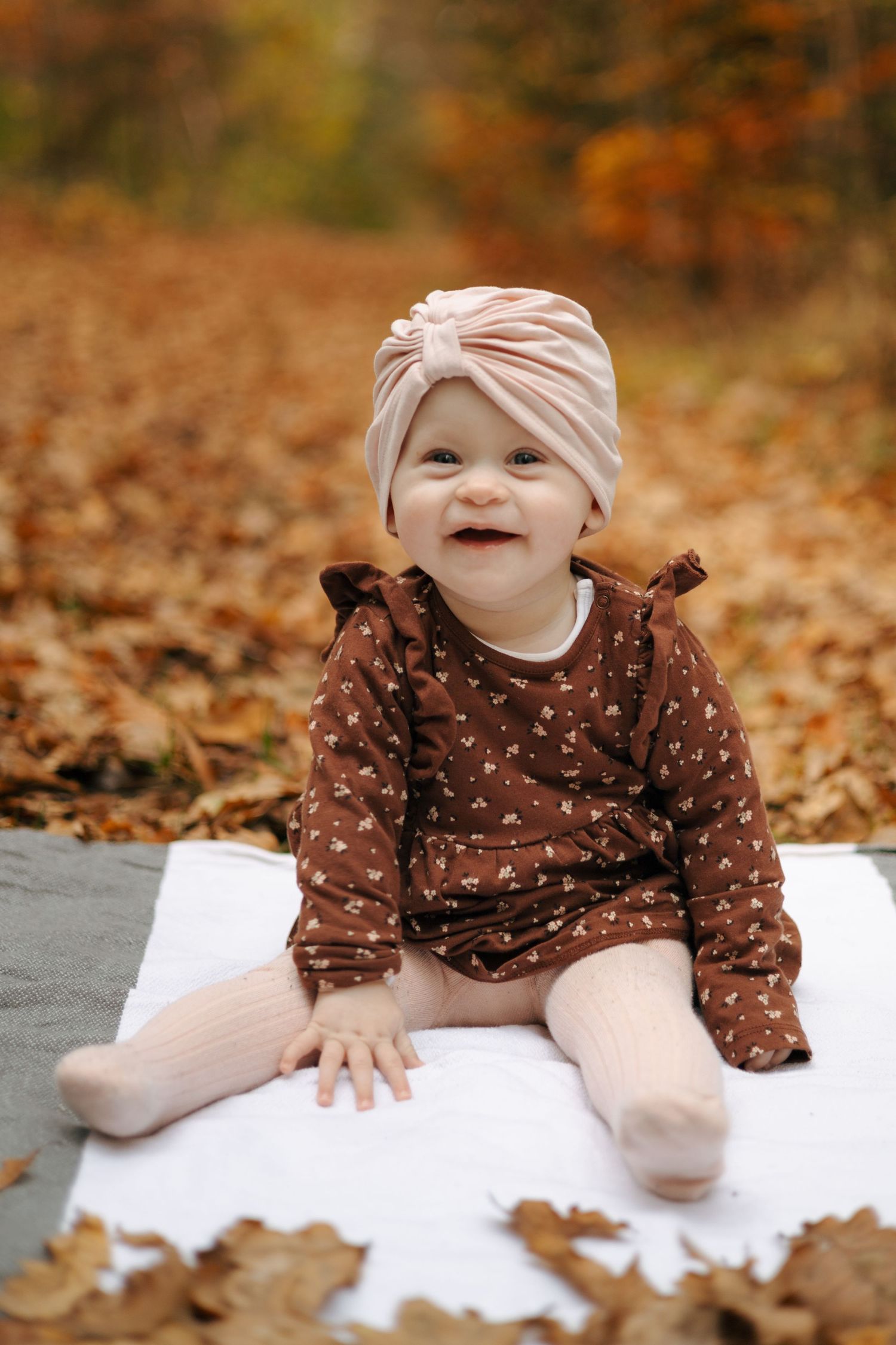 Baby in pink turban and brown dress sitting on white blanket surrounded by autumn leaves.