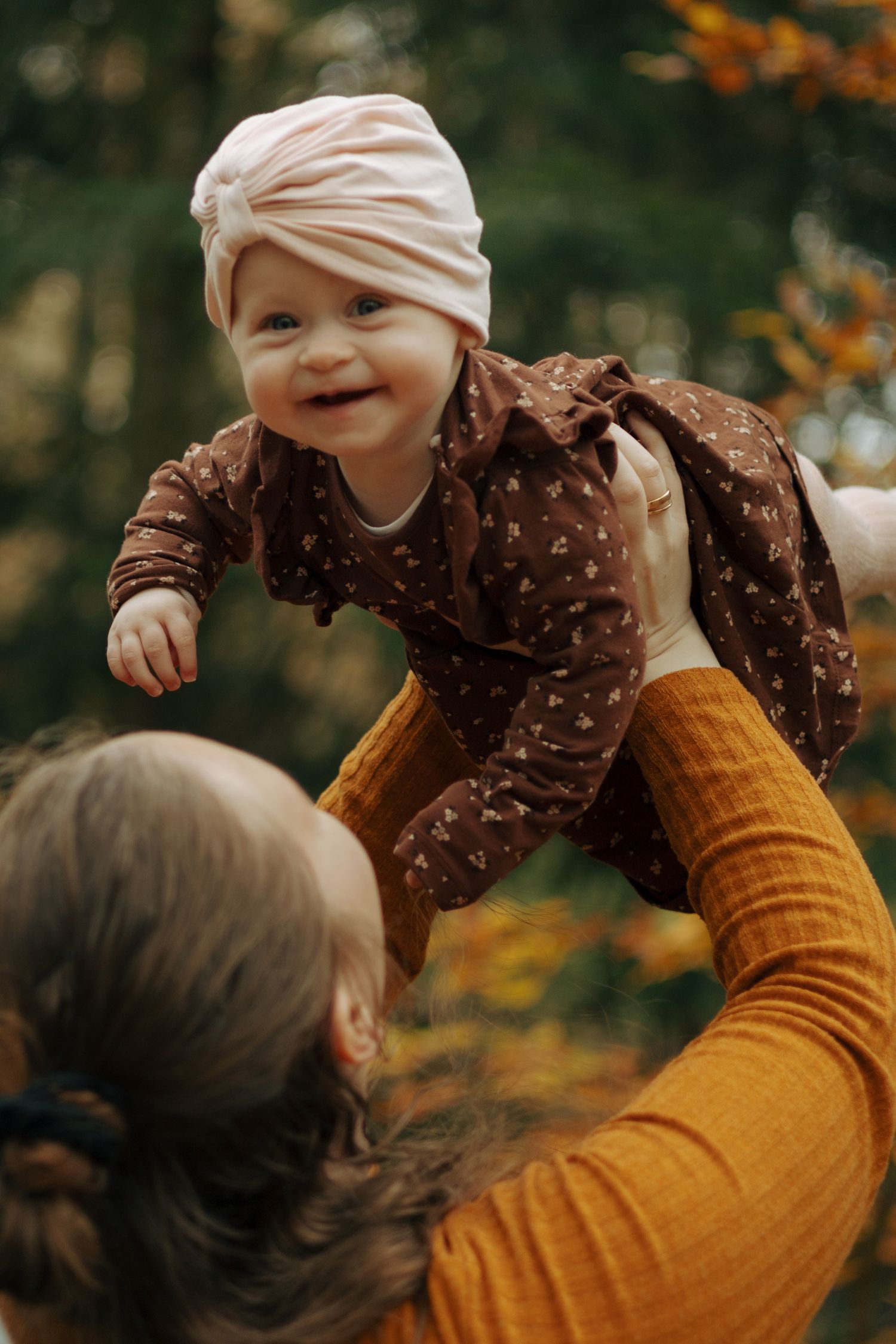 Baby wearing turban and polka dot dress being lifted up against autumn foliage background.