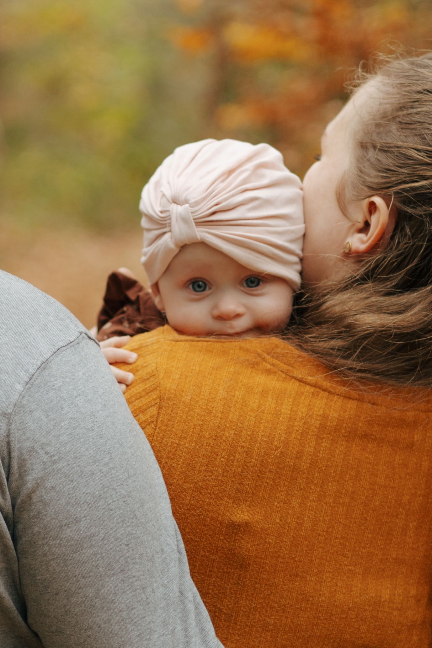 Baby wearing a pink turban peeks over shoulder against autumn background.
