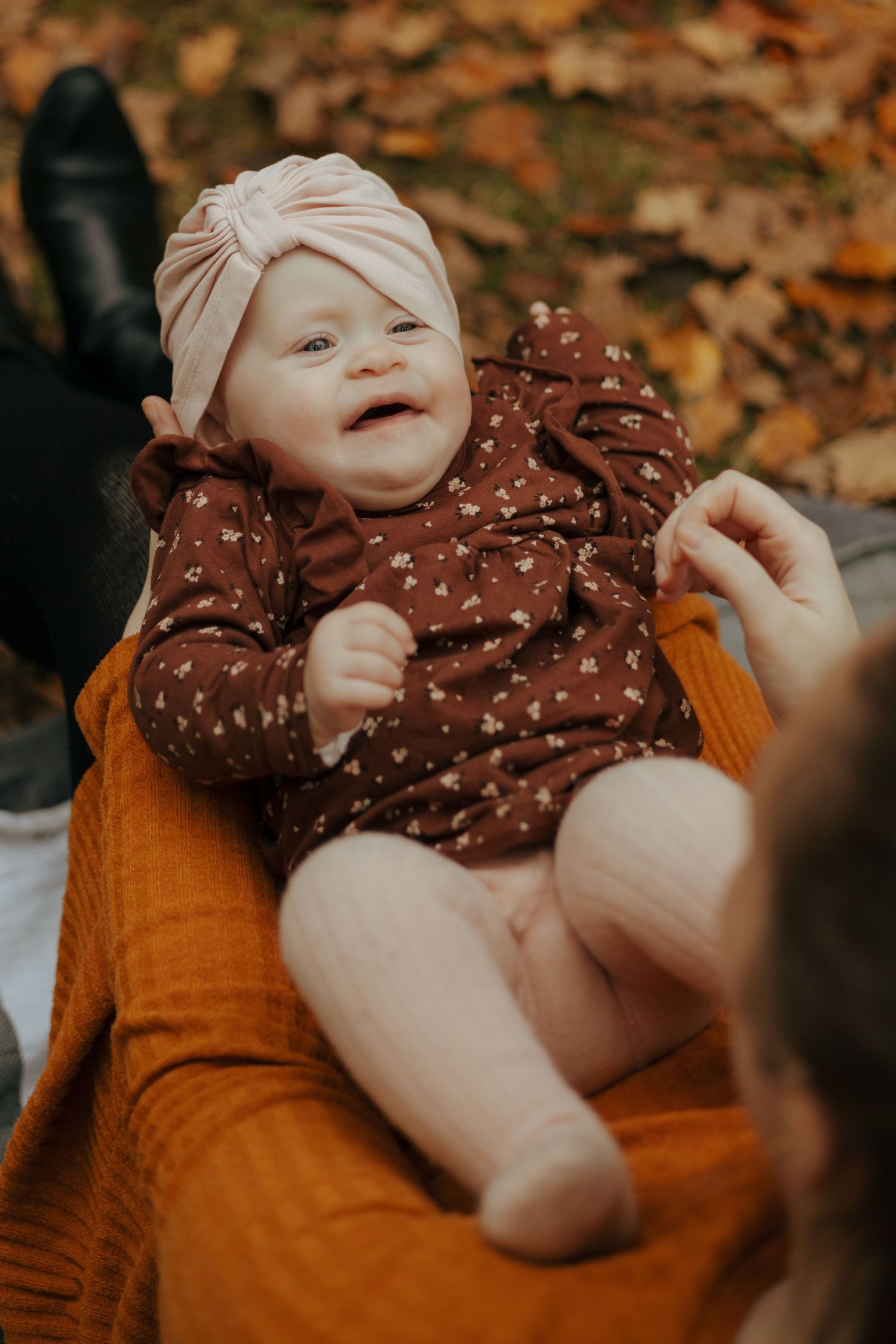 Happy baby in turban and polka dot dress sitting on parent's lap surrounded by autumn leaves.
