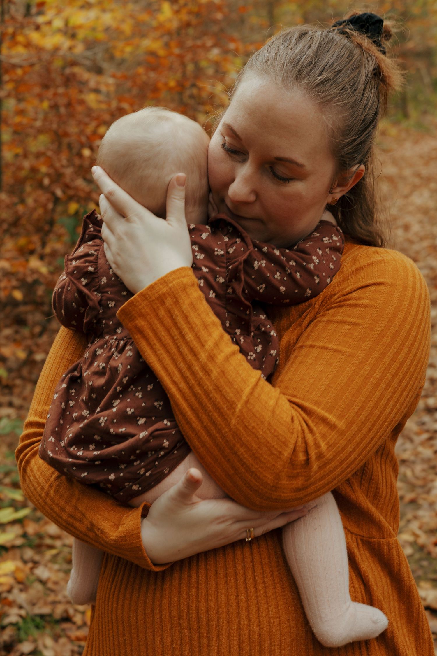 Parent in orange sweater holding baby in brown polka dot dress among fall foliage.