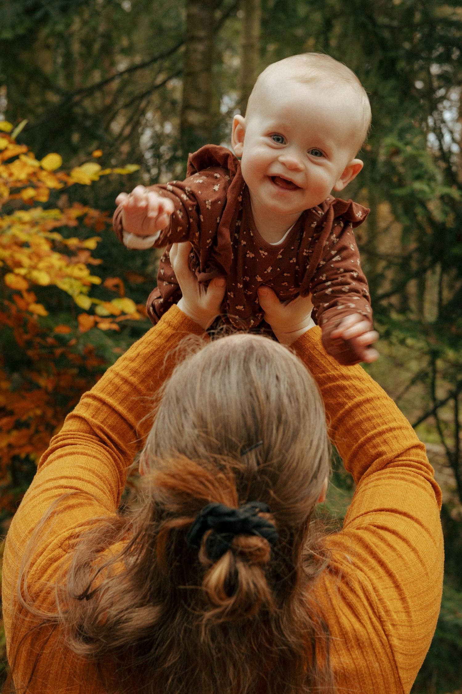 Joyful baby being lifted high in air against backdrop of autumn trees.