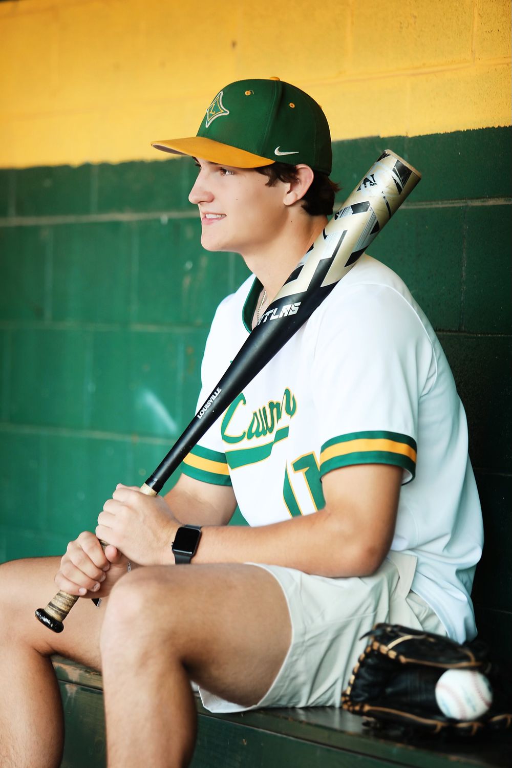 Baseball player in white Laurens uniform sits in dugout holding bat during game.
