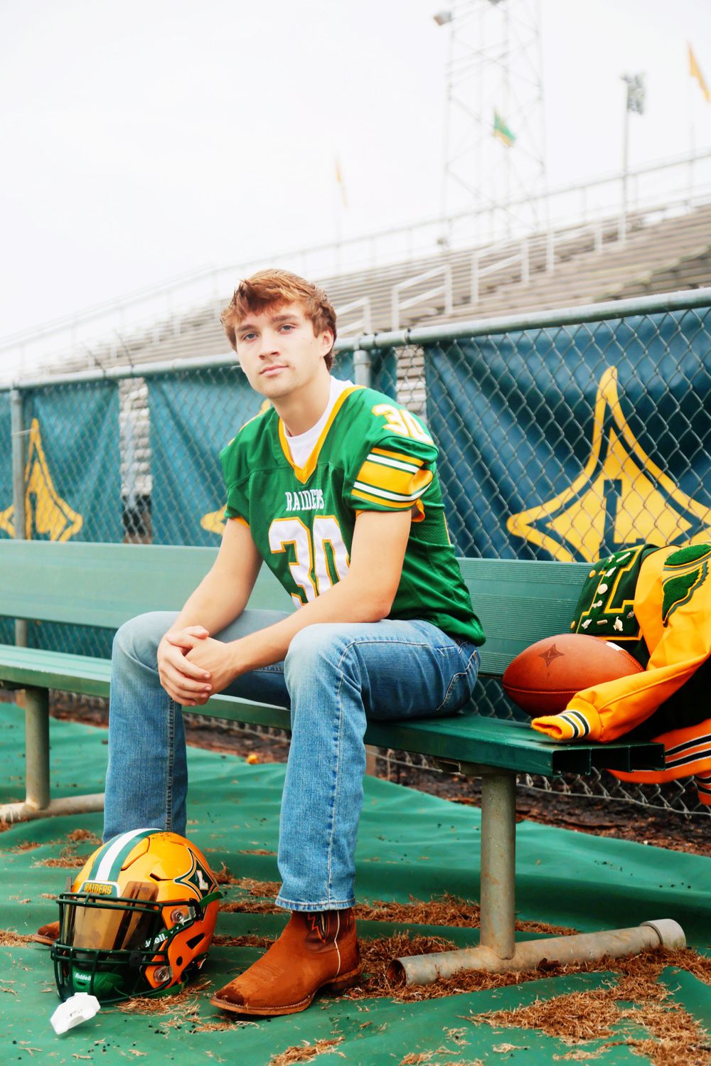 Football player sits on green bench with equipment and helmet nearby.