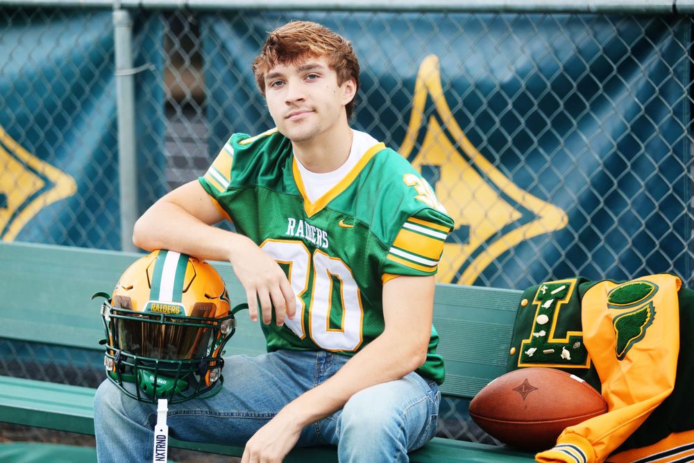 Football player in green jersey poses with helmet on stadium bench.