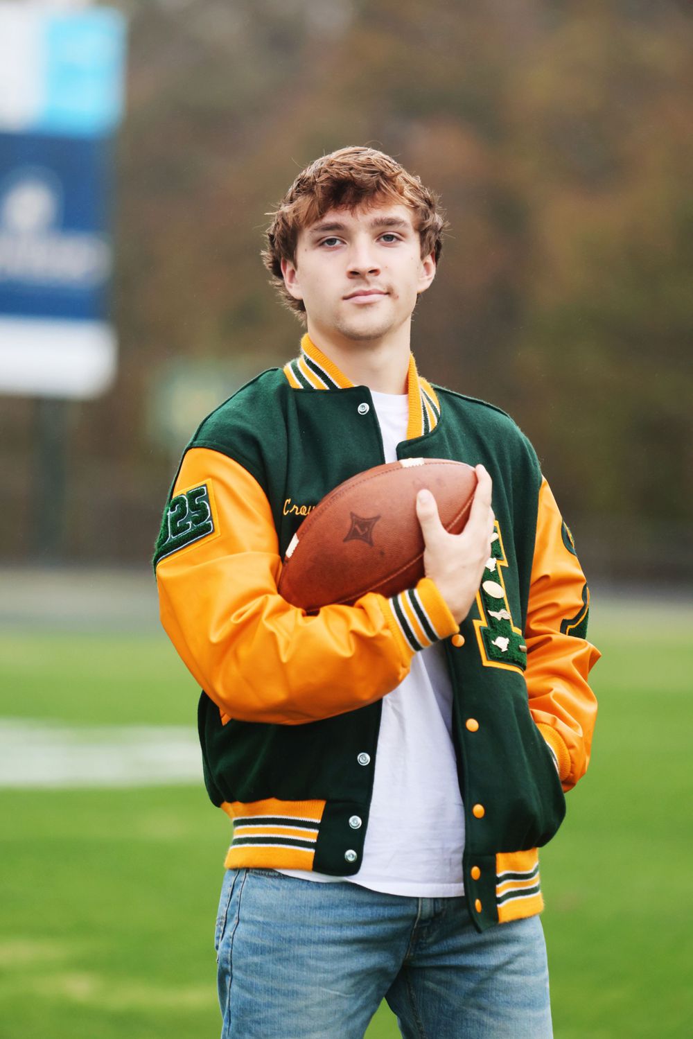 Football player wearing green and yellow varsity jacket holds ball on field.