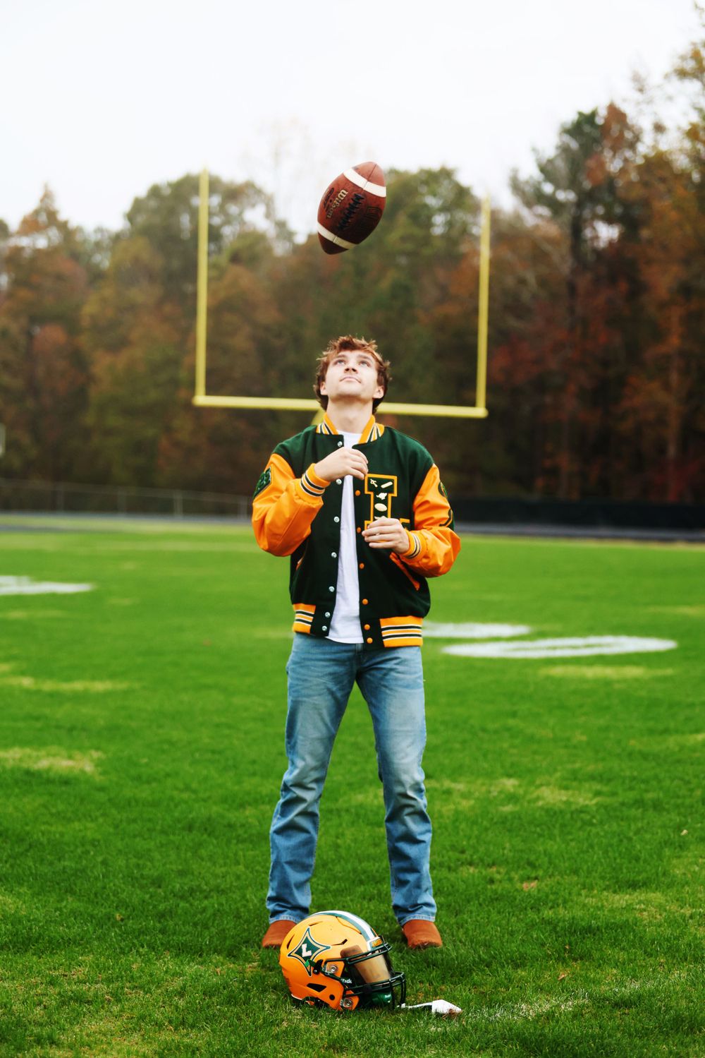 High school football player tosses football in air while standing on field with helmet at feet.