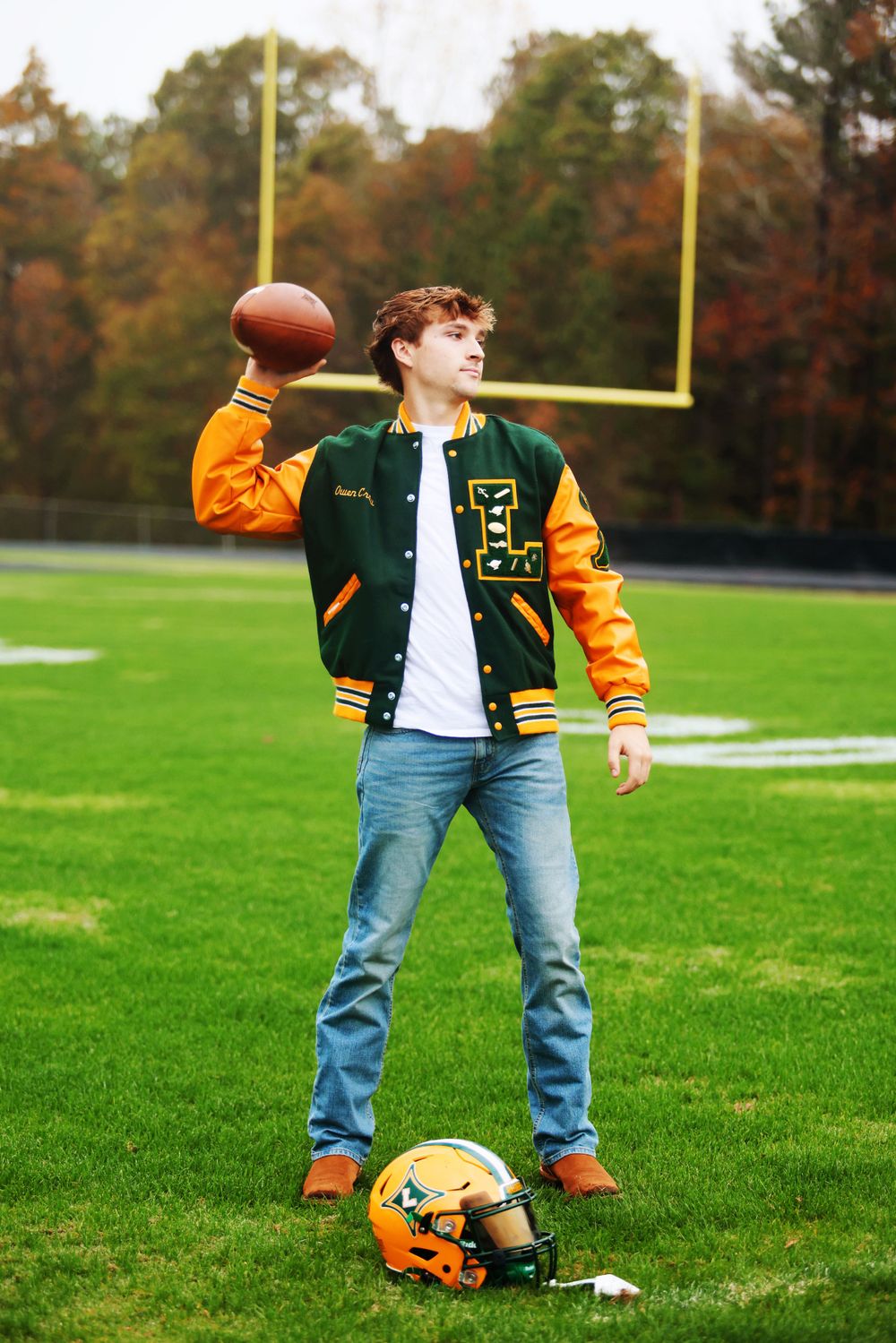Football player wearing varsity jacket throws football on field with goal posts in background.