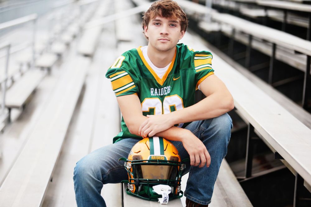 High school athlete sits on bleachers wearing green football jersey and holding helmet.