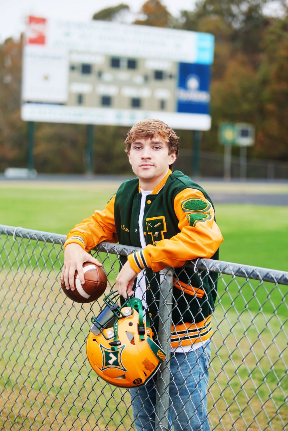 Student athlete poses with football and helmet against fence at school stadium.