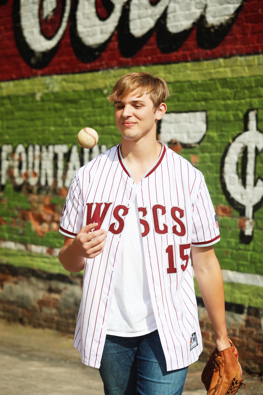 Baseball player poses with ball and glove near vintage Coca-Cola wall mural.
