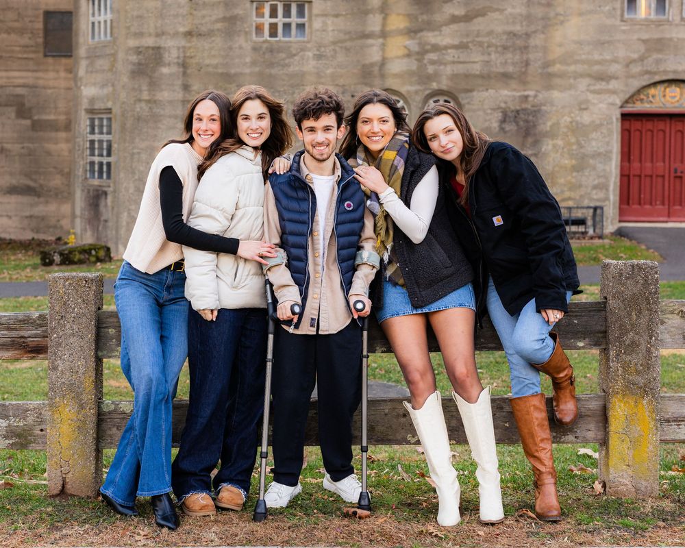 Six young friends smiling together outdoors near a historic stone building, one using crutches.