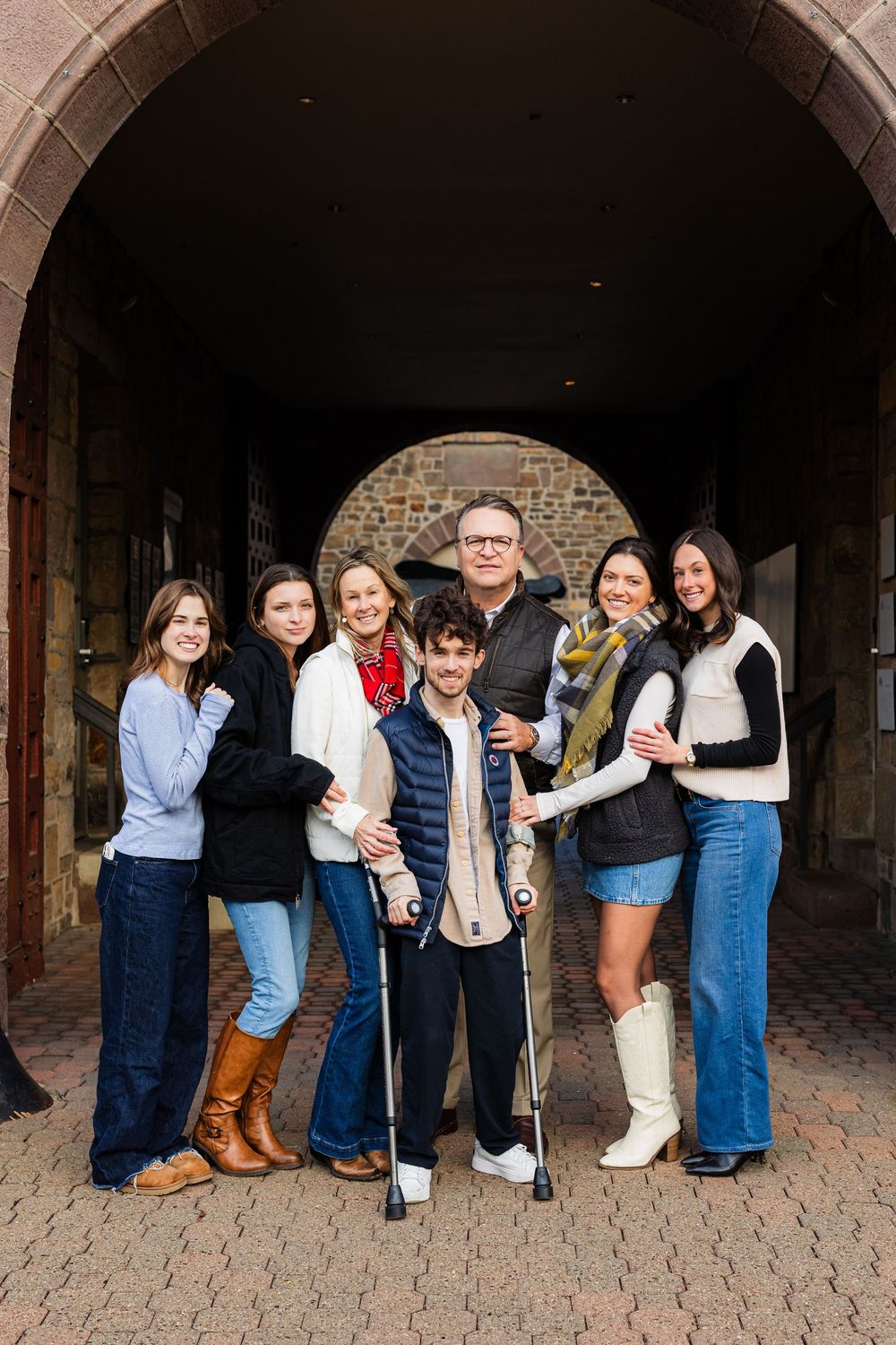 A family of seven poses together under a stone archway, one young man using crutches, all smiling warmly.