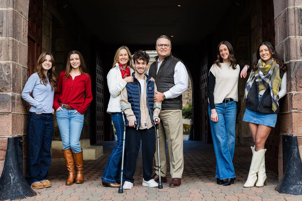 Family group of seven people smiling together in front of a brick archway, one member using forearm crutches.