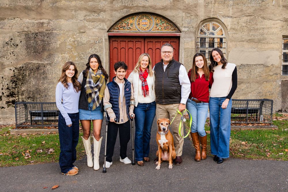 Family group of seven adults and a dog posing outdoors in front of a red door on a stone building.