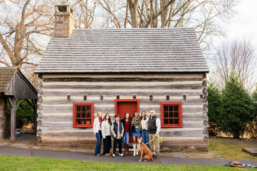 Group of people and a dog posing in front of a rustic log cabin with red-trimmed windows on a cloudy day.