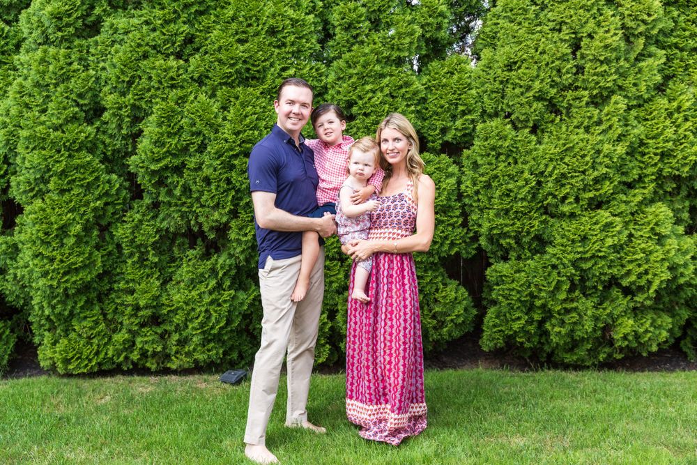A family group stands together on grass against an evergreen hedge backdrop.