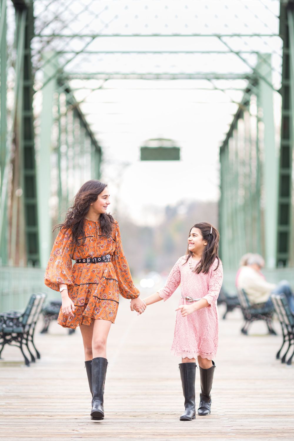 Two people walking hand in hand on a bridge wearing matching boots and dresses.