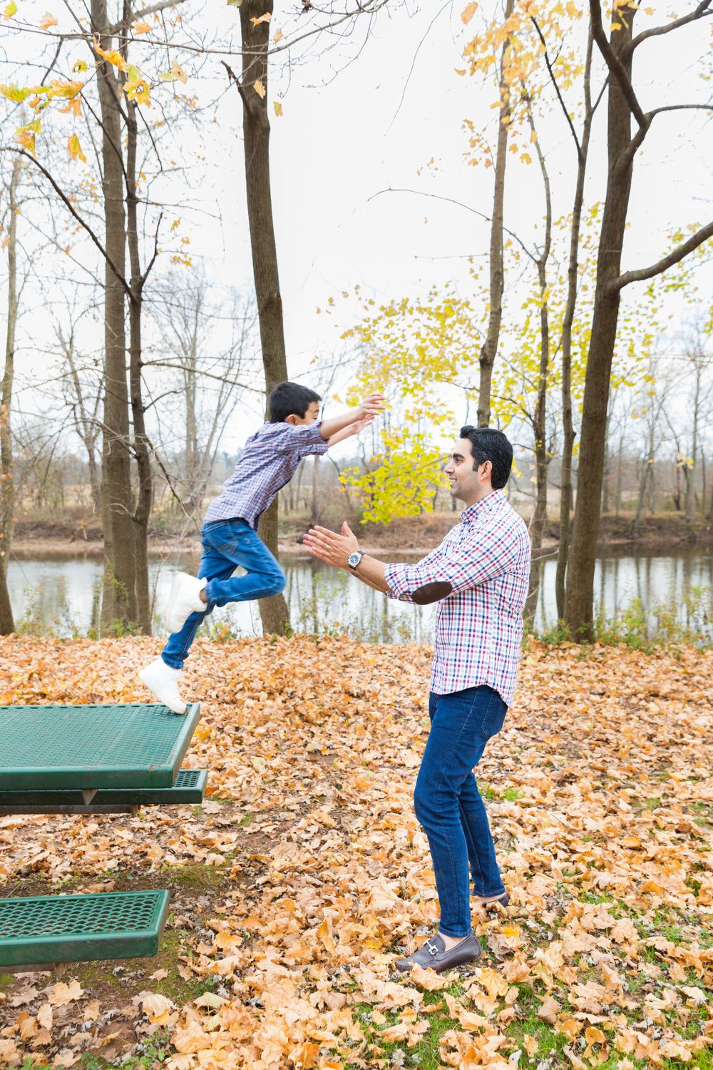 Two people playing and jumping on picnic tables covered in fall leaves near a lake.