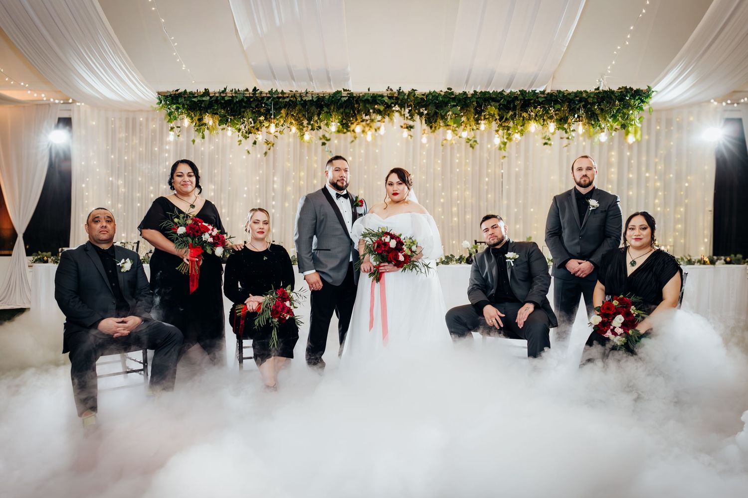 Wedding party poses for formal photo with smoke effects on white dance floor under greenery and lights.