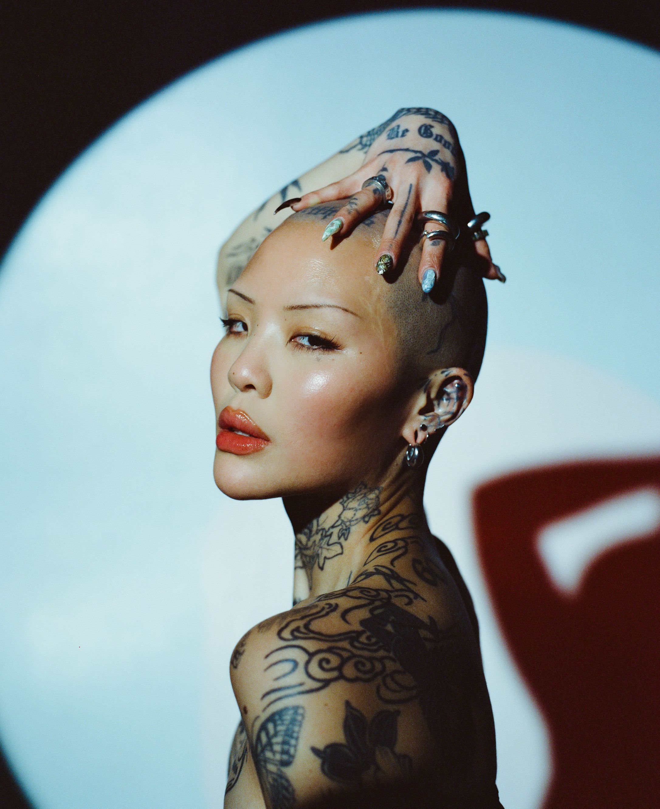 Artistic portrait of model with platinum hair, tattoos, and red lips against blue circular background.