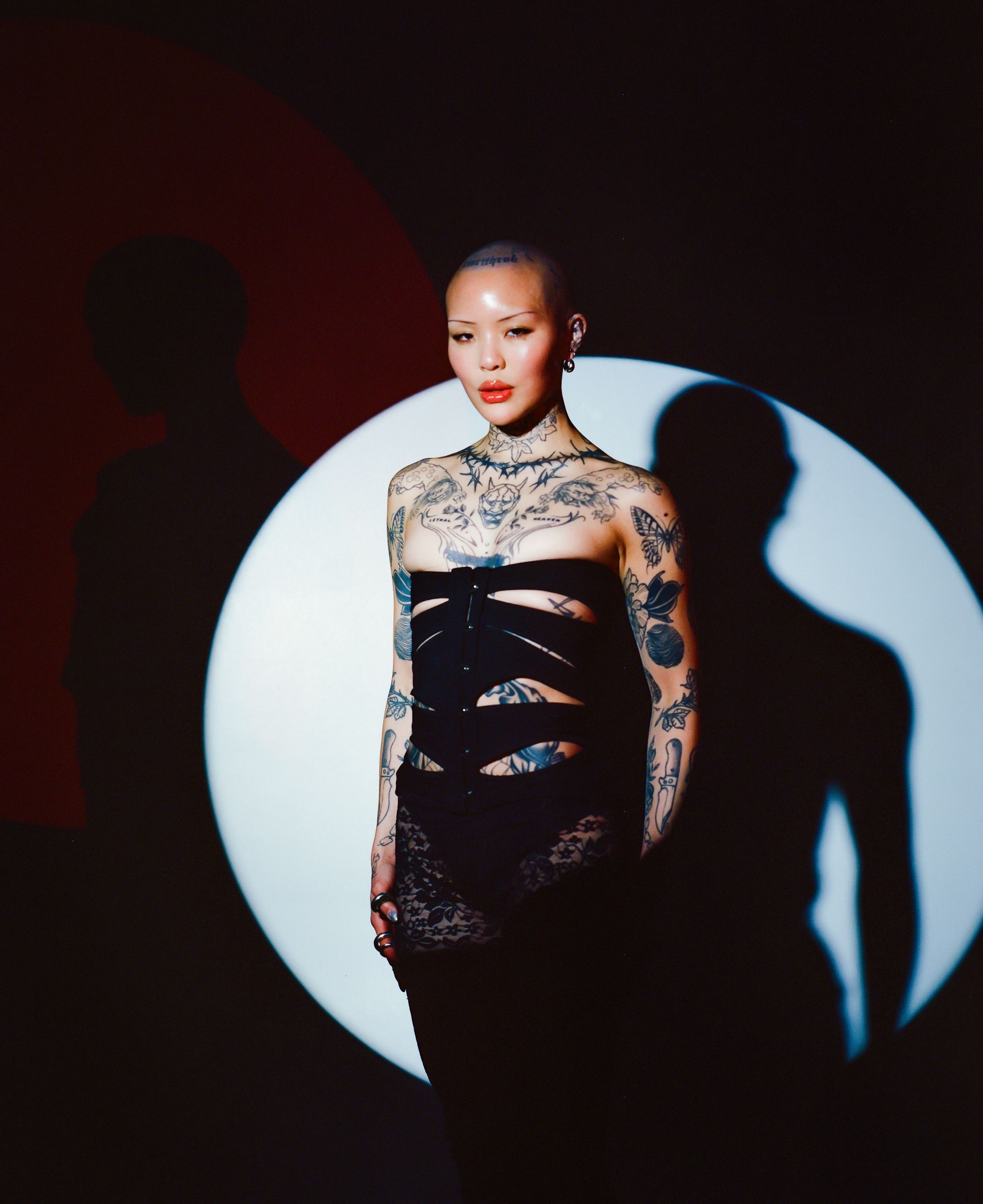 Model in black dress with tattoos posed against circular light backdrop.