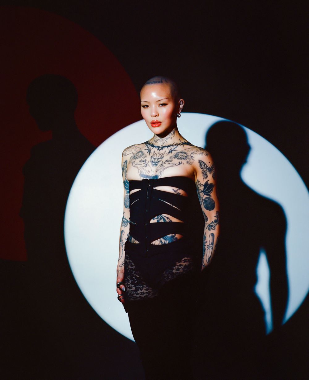 Model in black dress with tattoos posed against circular light backdrop.