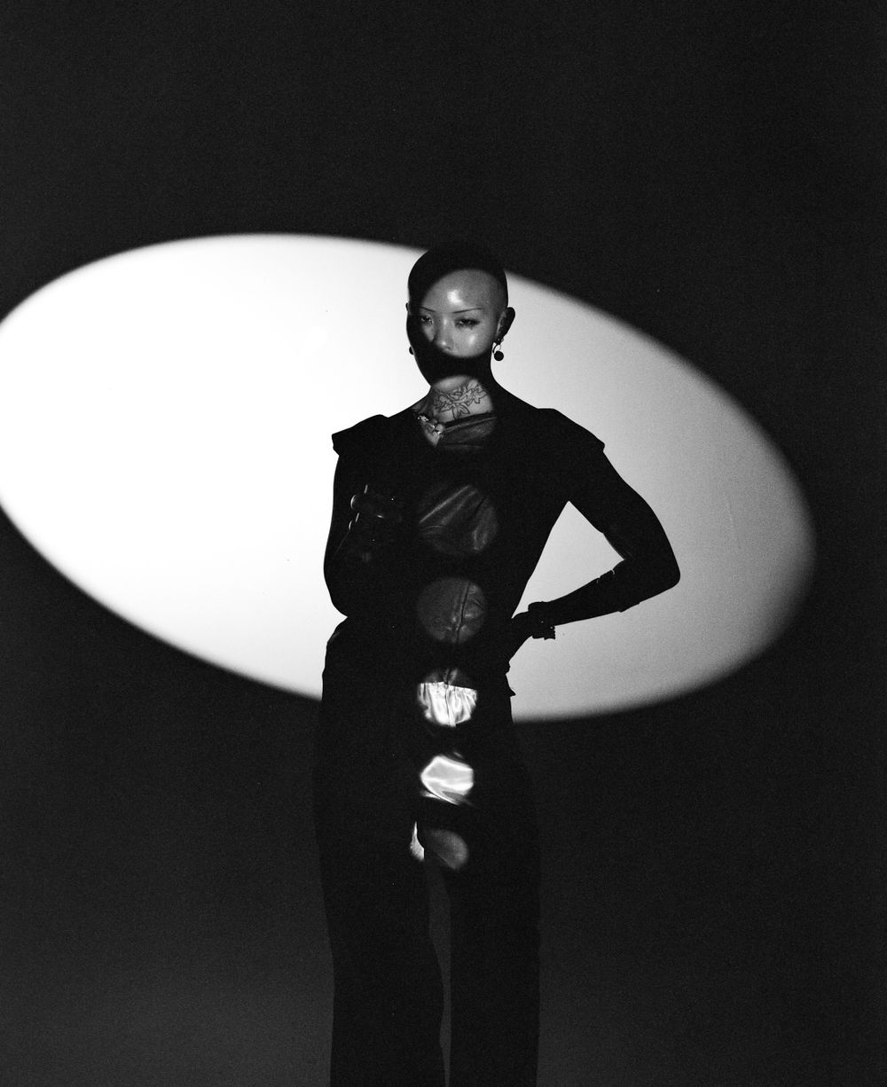 Dramatic black and white fashion portrait with model in dark outfit against oval spotlight backdrop.