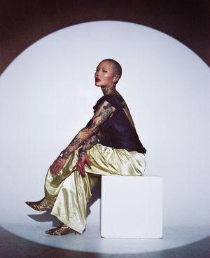 Model seated on white cube wearing gold pants and dark top against circular backdrop.