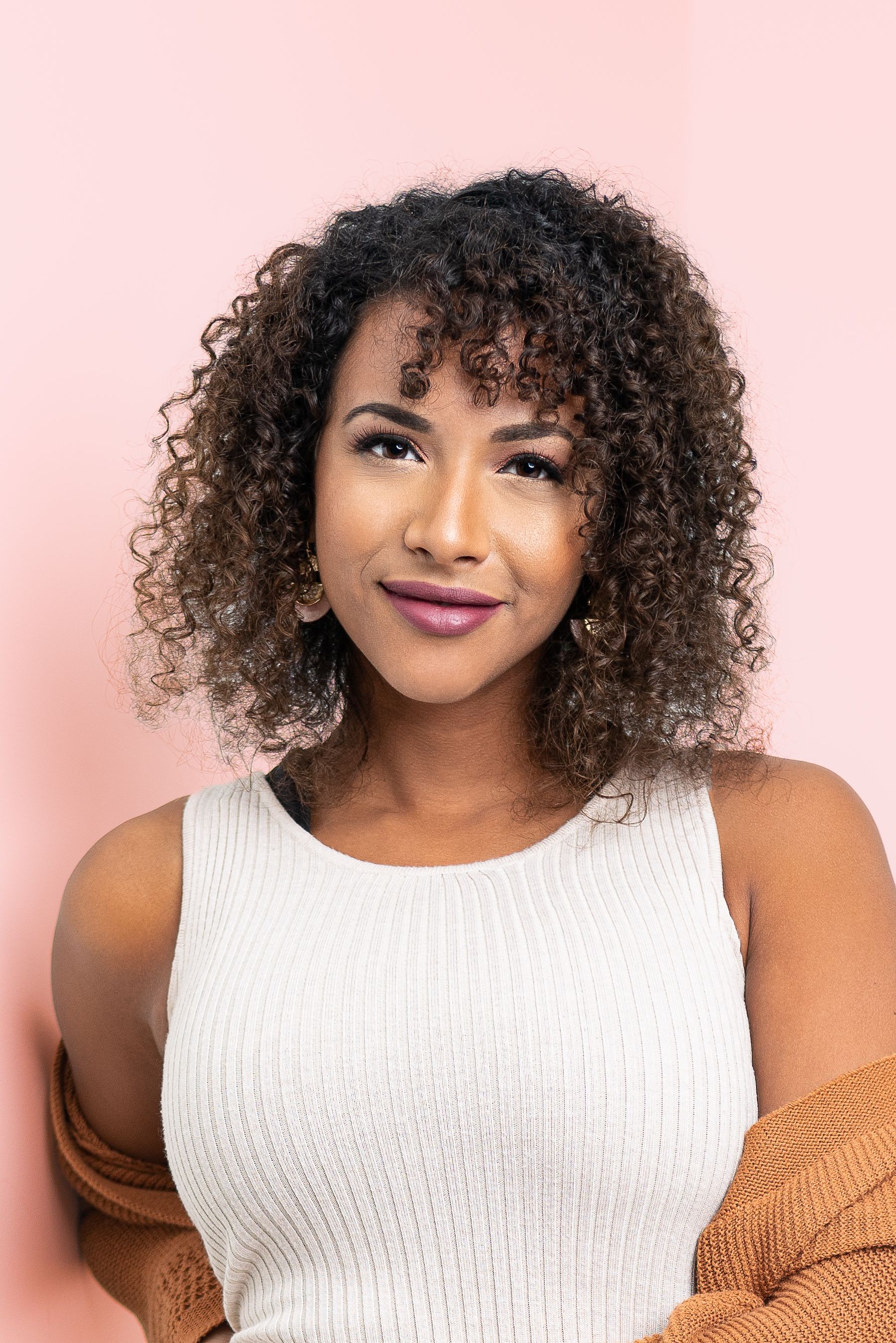 Portrait of someone with curly hair wearing a white tank top and orange cardigan against a pink background.