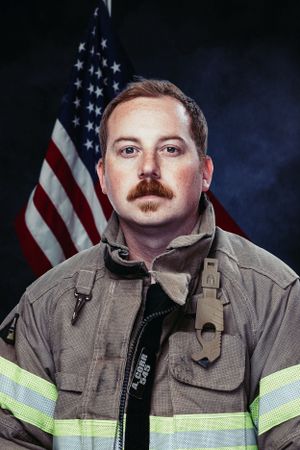 Firefighter in protective gear posing for official portrait with American flag backdrop.