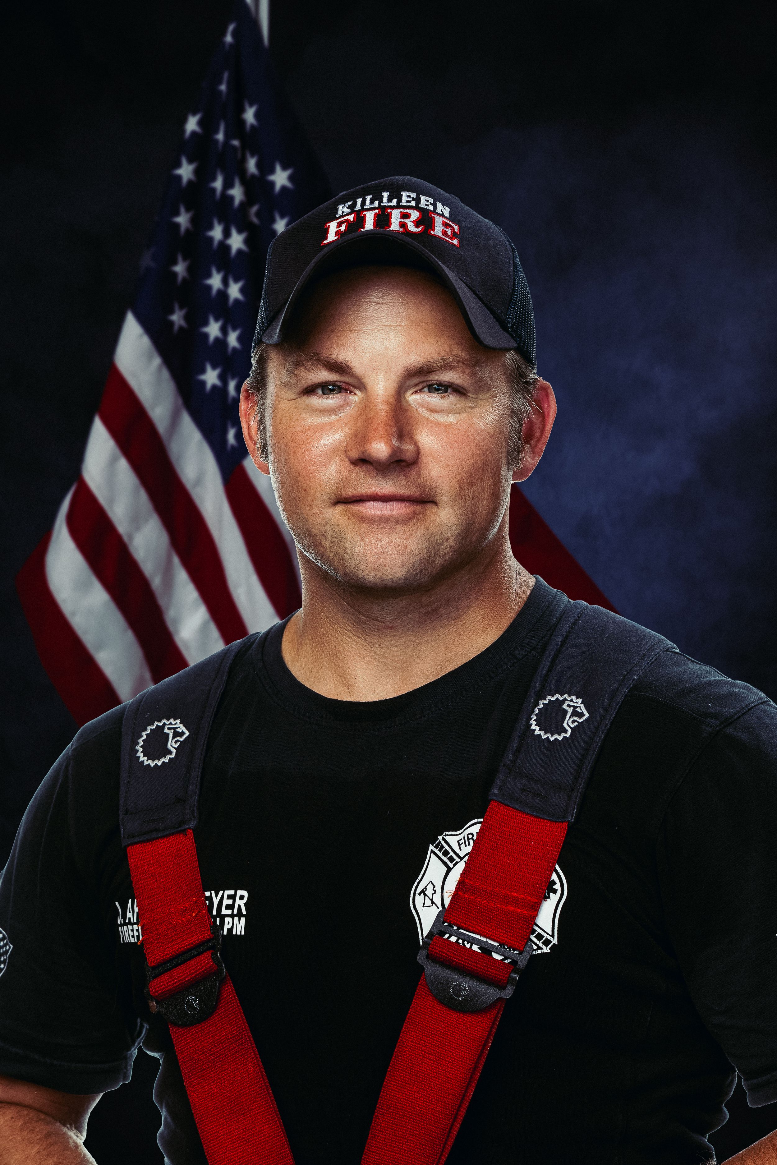 Firefighter in black t-shirt and red suspenders posing in front of American flag.