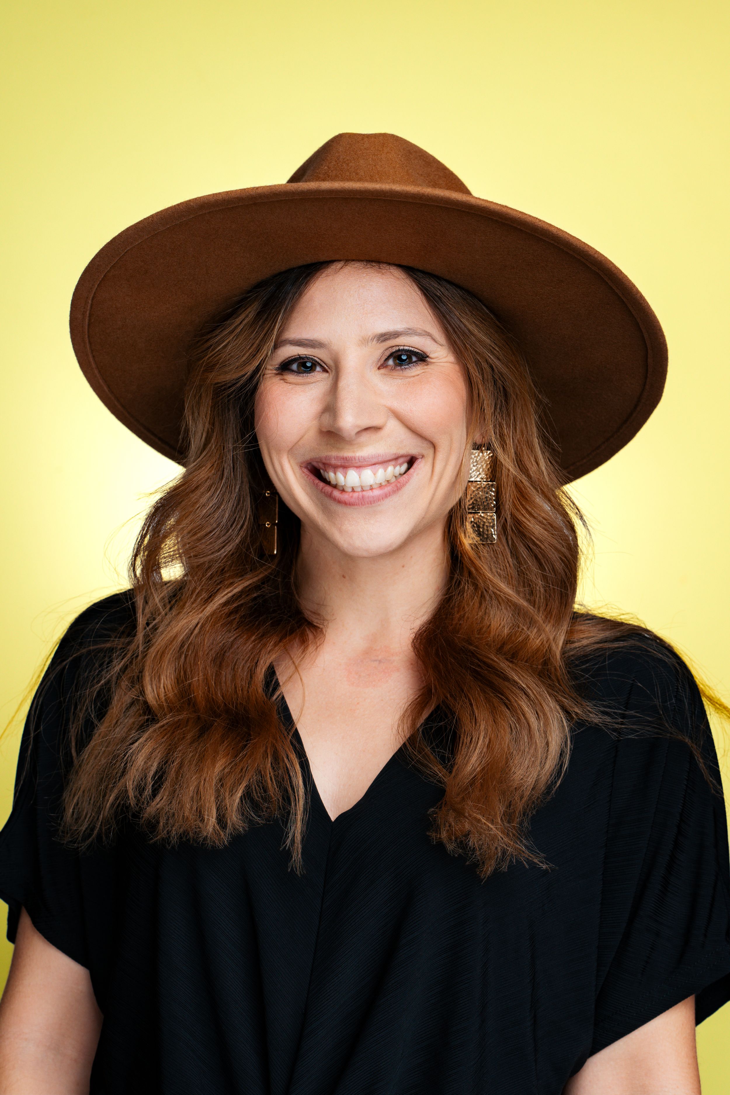Professional headshot of a person in a brown hat and black top against a yellow background smiling warmly.