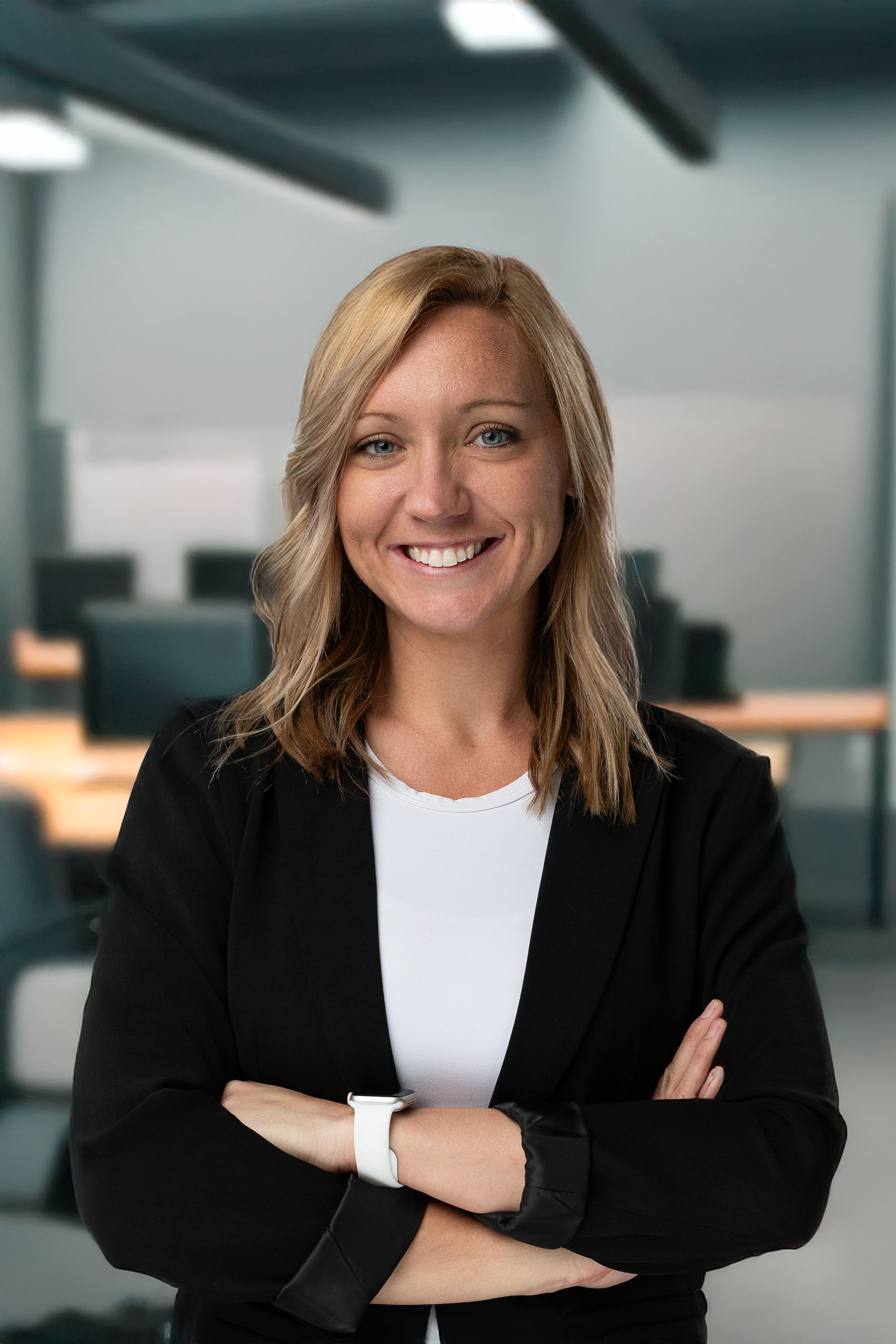 Professional portrait of person in black blazer and white top with arms crossed in office setting.