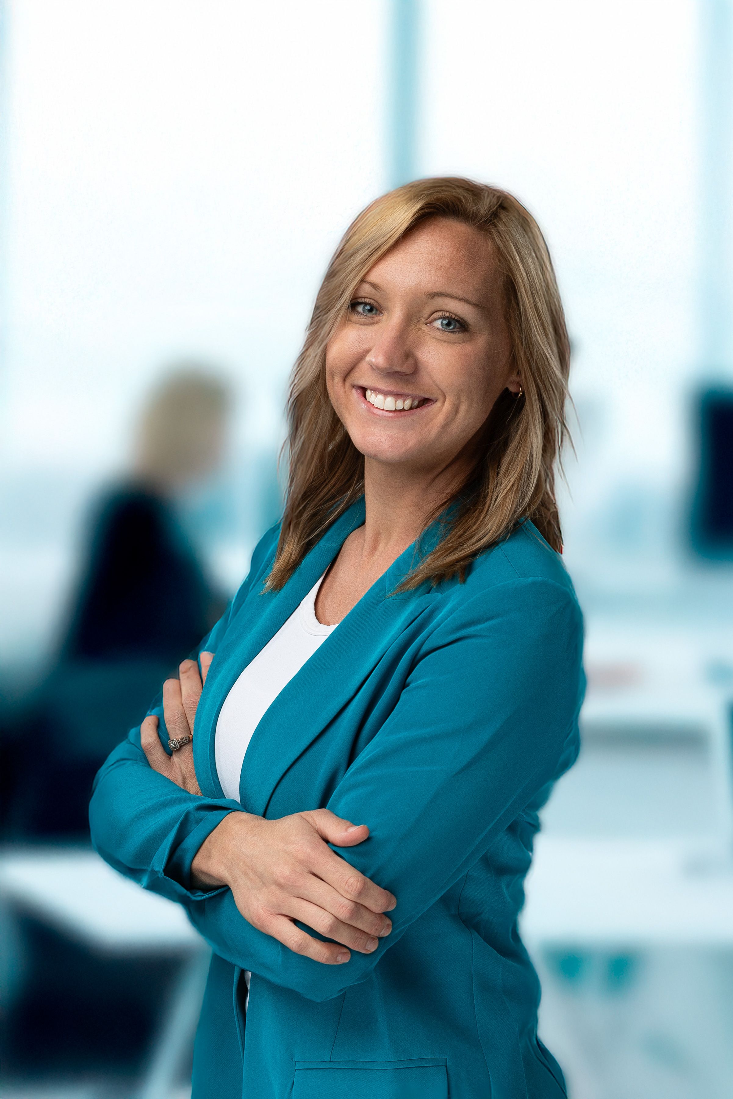 Professional portrait of person in teal blazer with crossed arms in office environment.