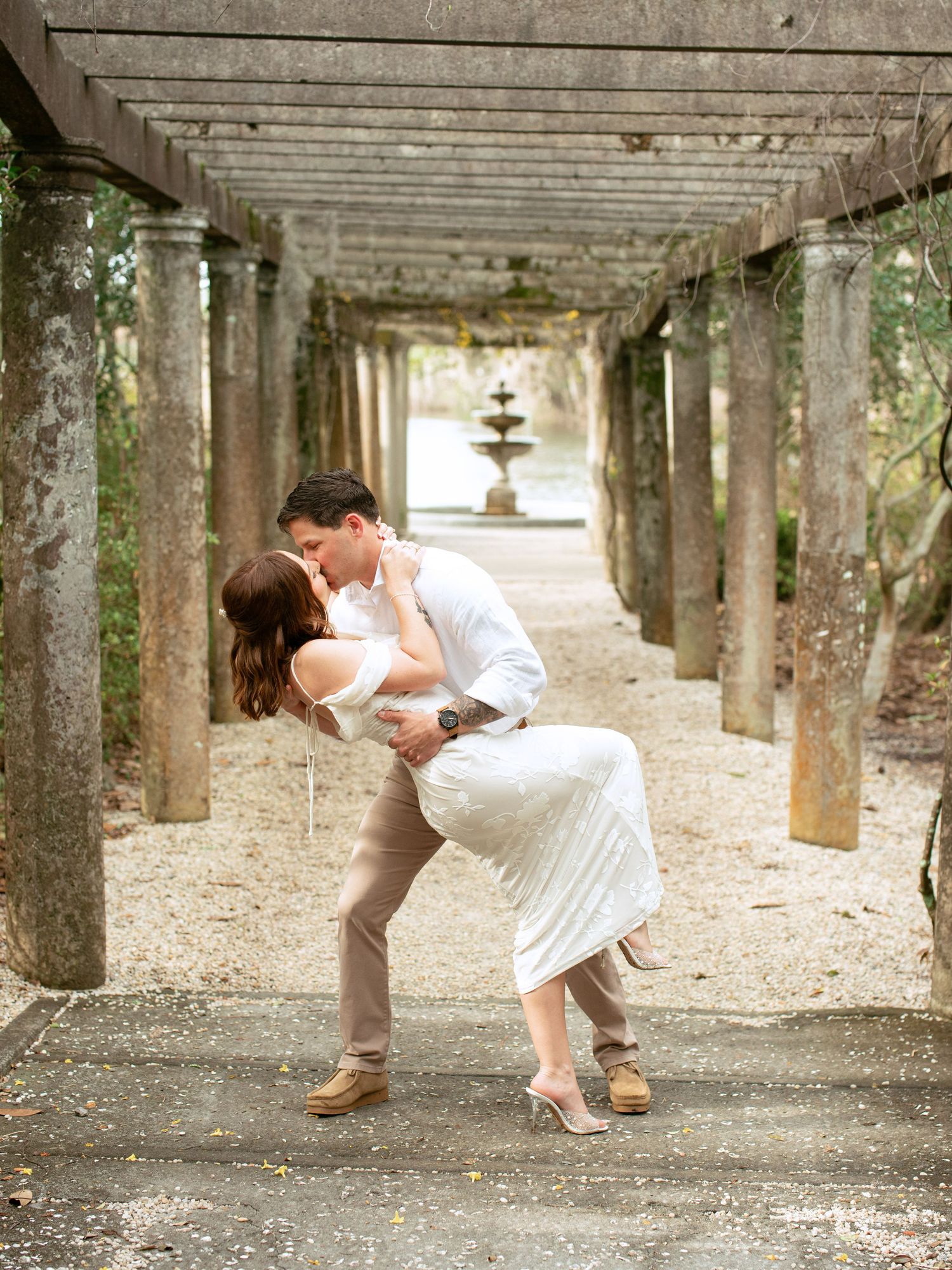 A romantic couple embraces under a rustic pergola with stone columns and a fountain visible in the distance.
