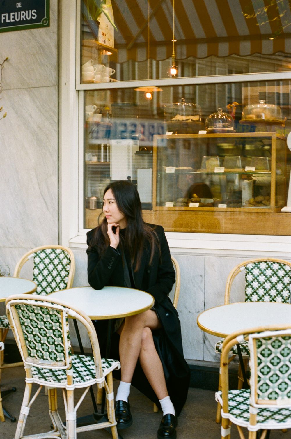 A person sits at an outdoor Parisian cafe with rattan chairs and round marble tables near a storefront window.