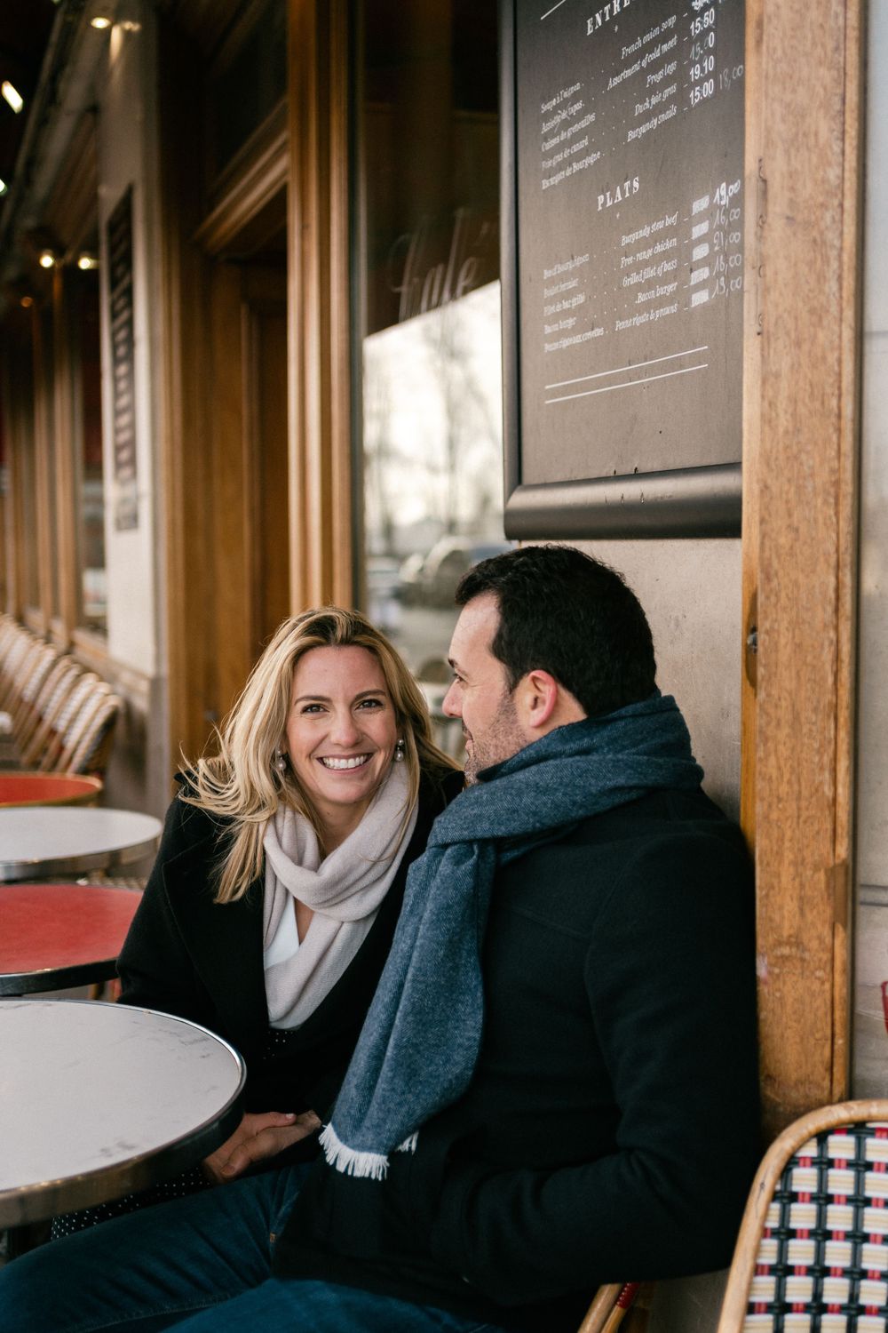A couple enjoying conversation at an outdoor Parisian cafe with round white tables and wooden exterior.