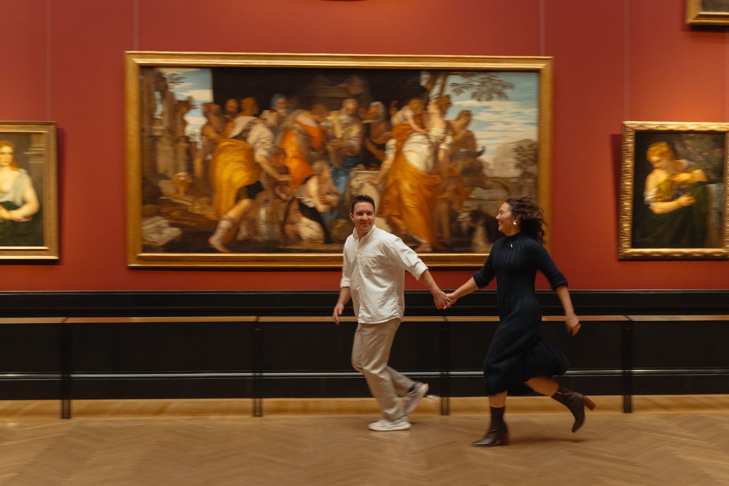 Two people run playfully through an art gallery with classical paintings displayed on red walls behind them.