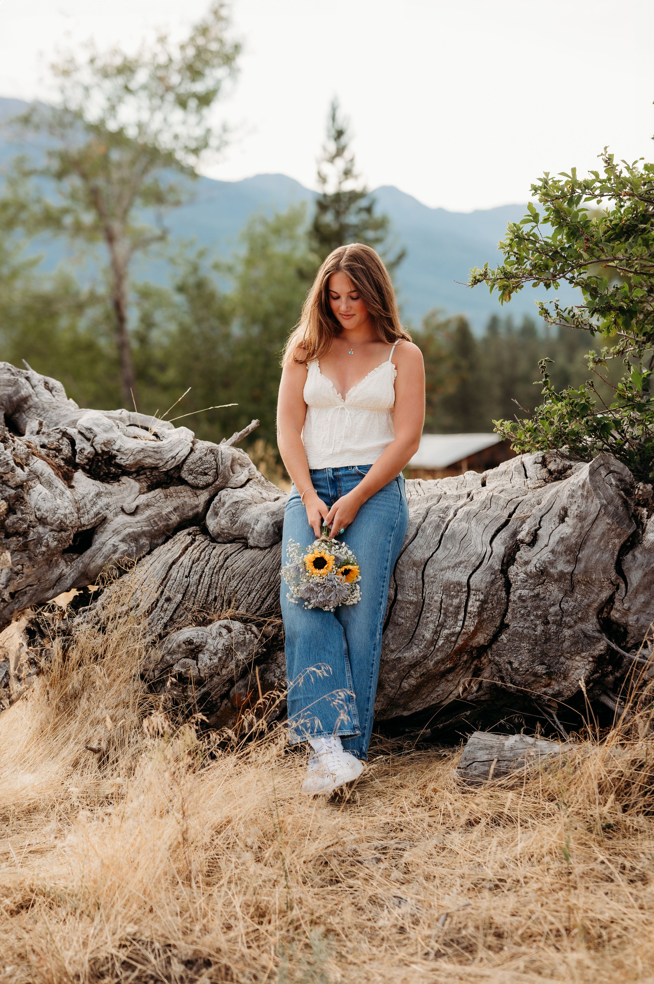 Figure in white top and jeans poses with sunflower on fallen log in mountain setting. Wallowa Lake, Joseph, Oregon