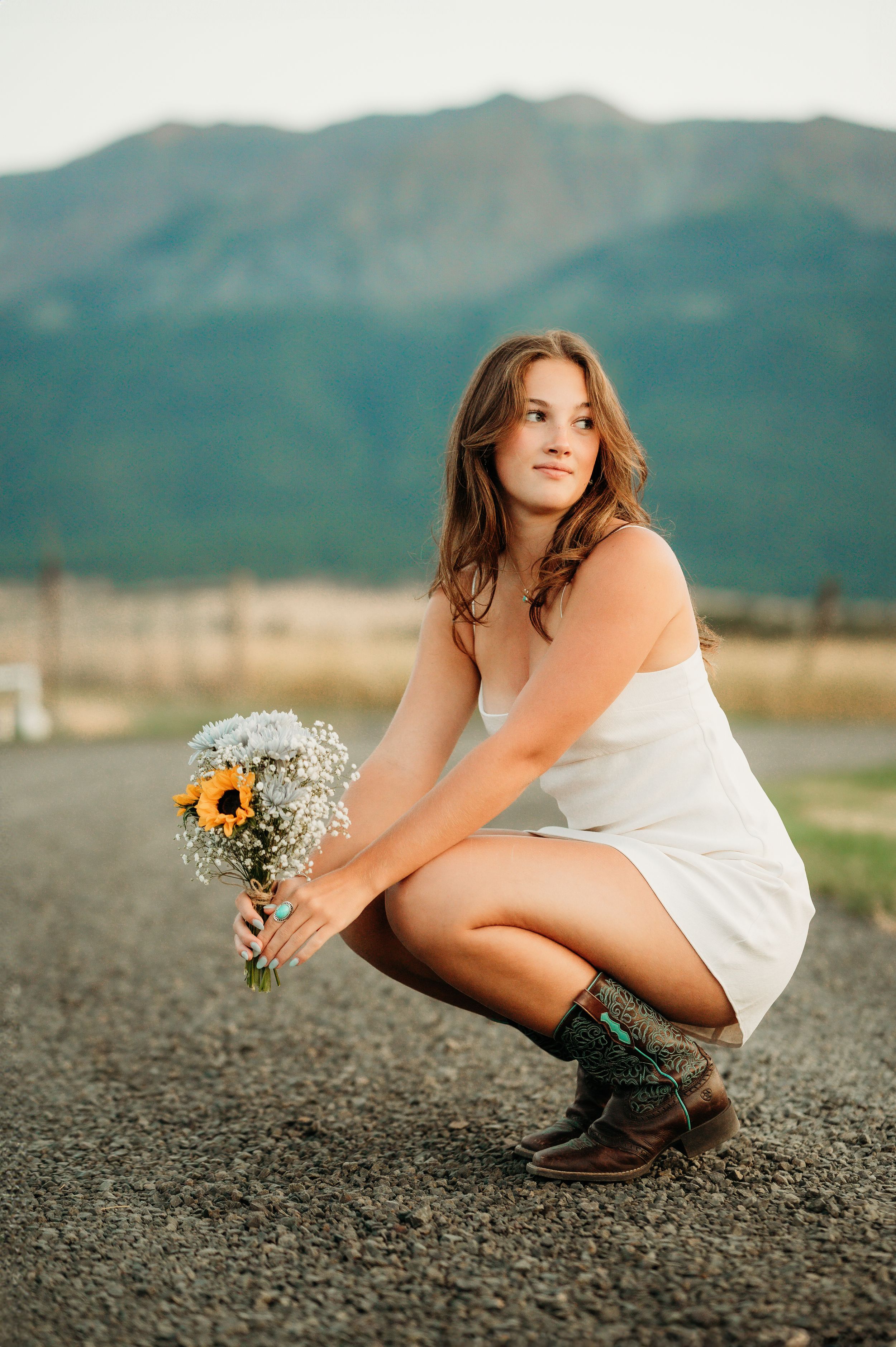 Figure in white dress crouches on dirt path holding wildflowers with mountains behind.