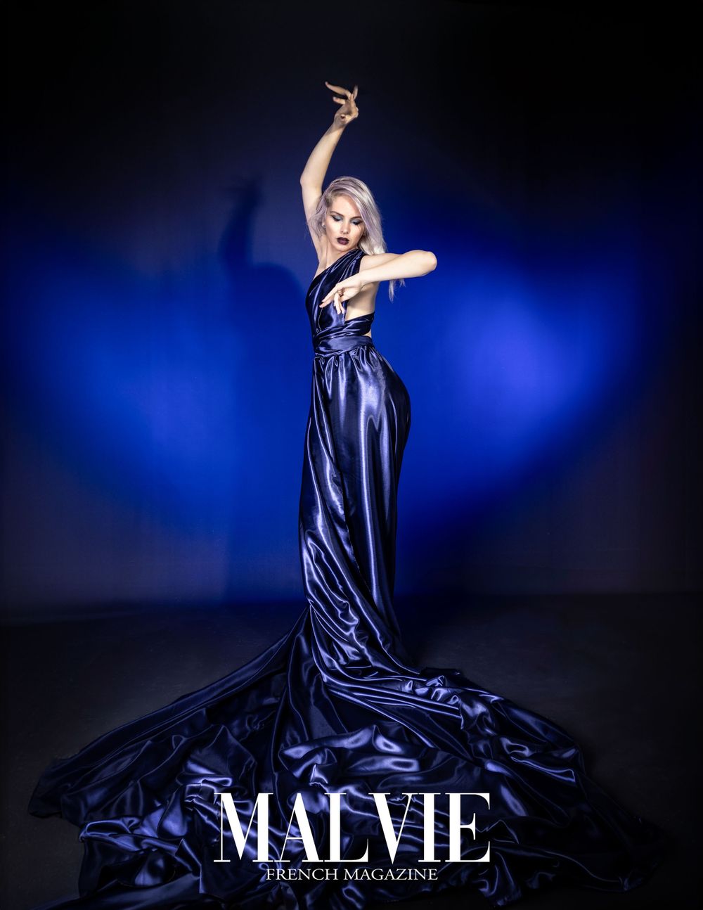 Elegant model in flowing navy blue satin gown poses dramatically against dark blue backdrop.