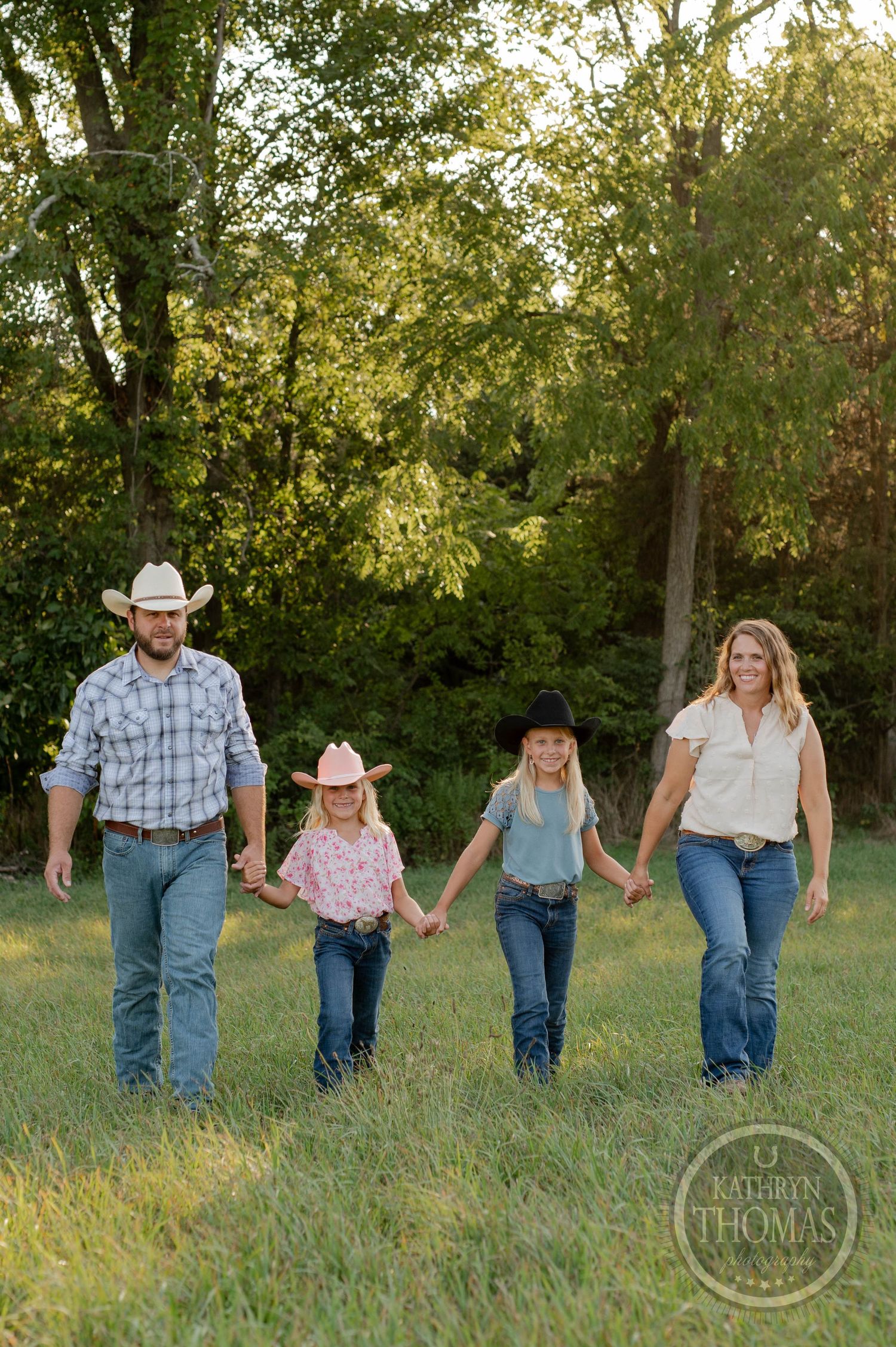 Family wearing casual western attire holds hands while walking through a green field.