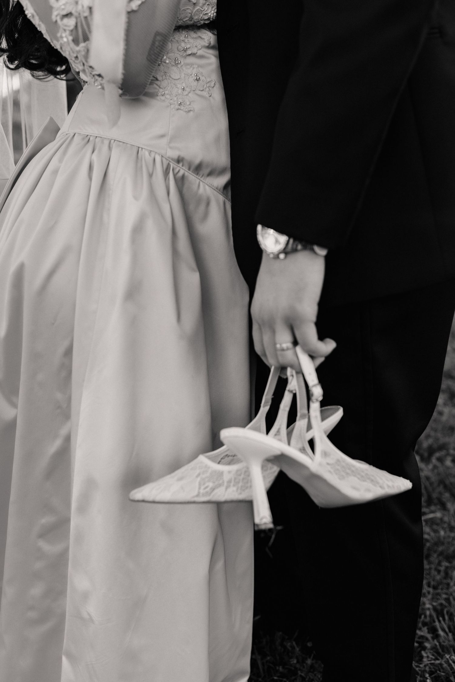 A black and white detail shot of formal wedding attire and held bridal shoes.