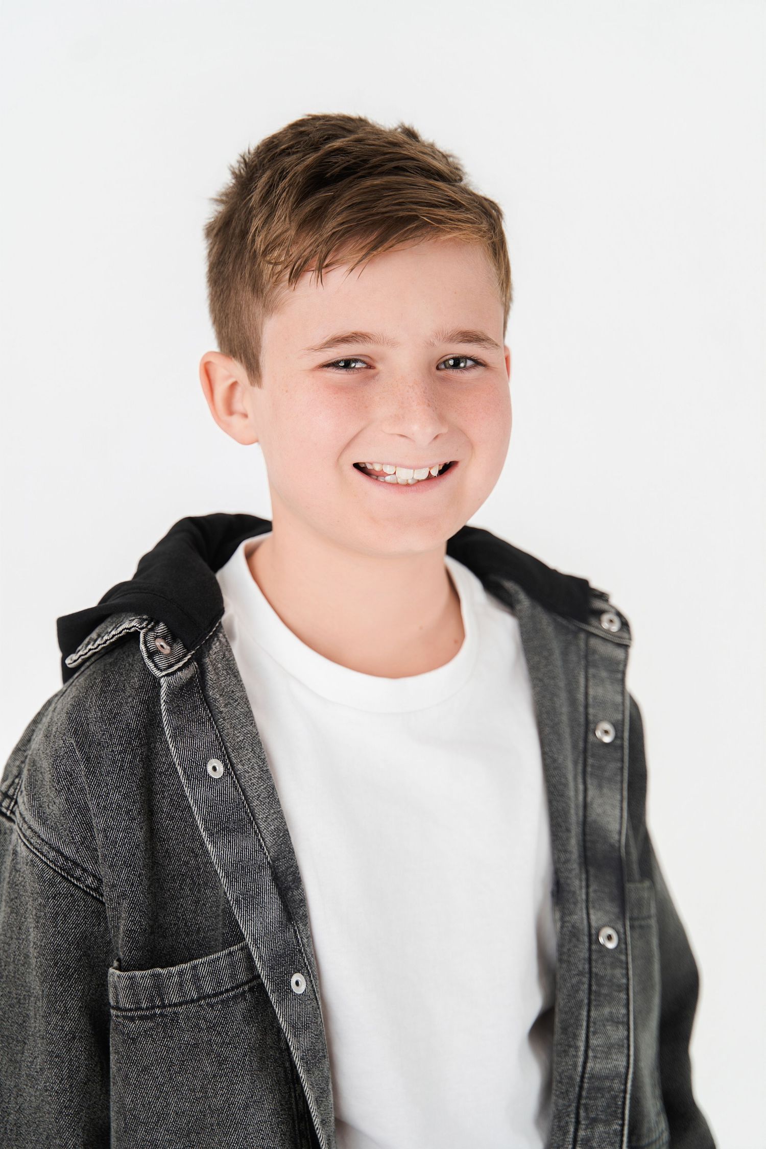 Young boy smiling wearing a black denim jacket and white t shirt, posing against a white background, studio actor headshot