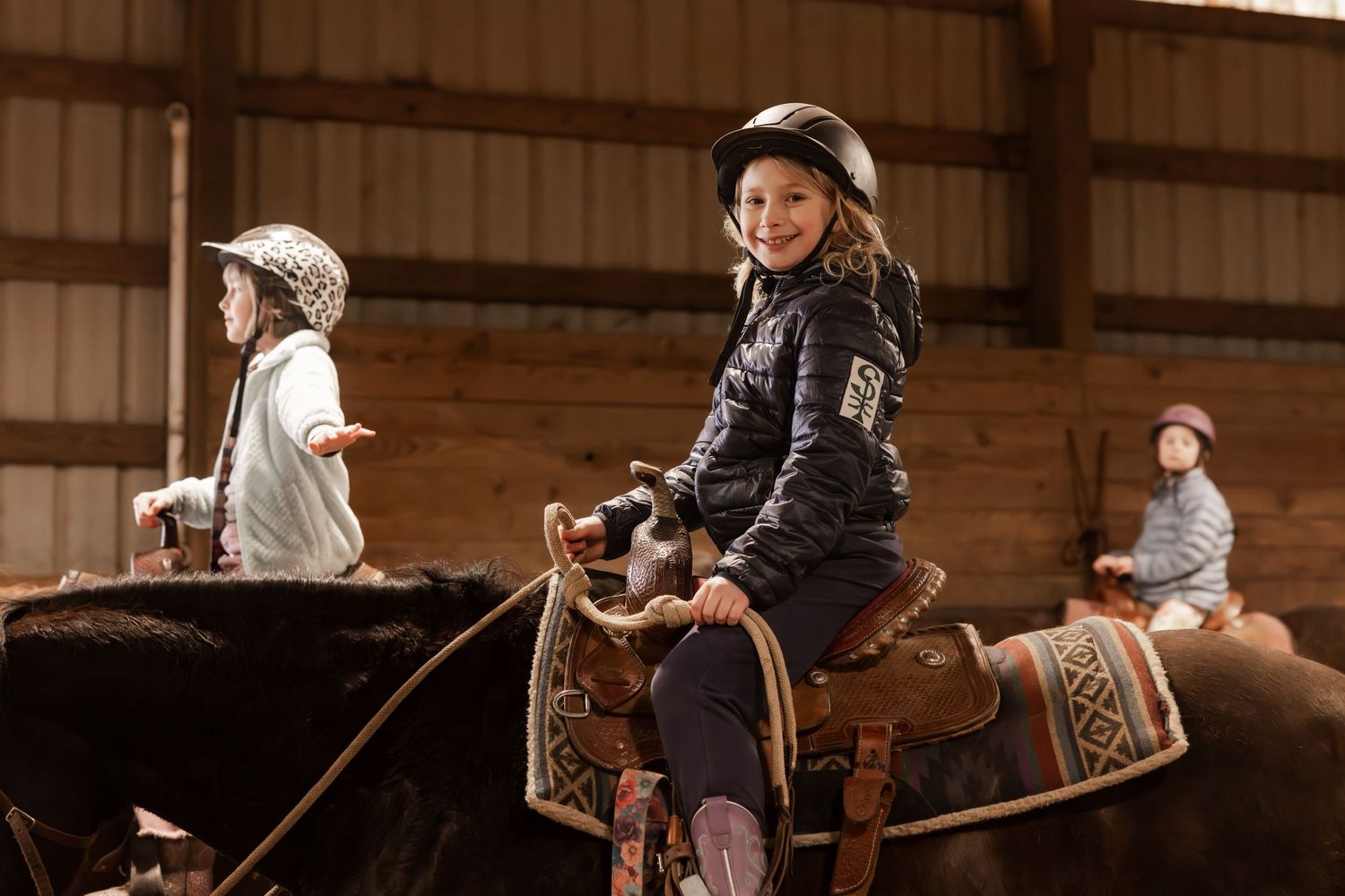 Young riders practicing horseback riding skills inside a wooden barn with western-style training equipment and saddles.
