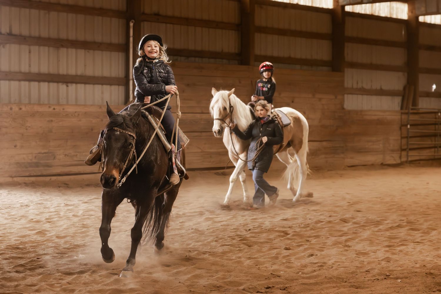 Riders practicing horseback riding inside a covered arena with sandy ground and wooden walls.